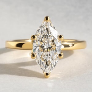 May include: A gold engagement ring with a marquise-cut diamond. The diamond is set in a four-prong setting and is the focal point of the ring. The band is a simple, polished gold. The ring is displayed on a white surface.