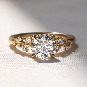 May include: A gold ring with a large, round, clear diamond center stone. Smaller diamonds are clustered on either side of the center stone. The ring is set against a white background.