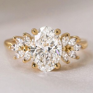 May include: A gold ring featuring a large, oval-cut diamond as the centerpiece. The ring is adorned with smaller, petal-shaped diamonds on either side of the main stone, creating a floral design. The band is a simple, polished gold.