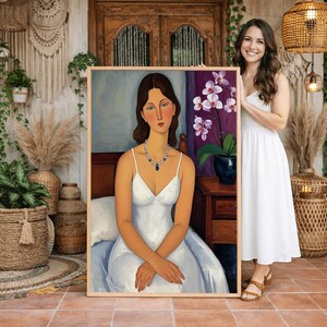 Puede incluir: Retrato enmarcado de una mujer con un vestido blanco, sentada en una cama, con un collar azul. La pintura tiene un marco marrón claro. El fondo incluye una cortina morada, una mesita de noche y una orquídea en maceta. La mujer tiene el pelo castaño.