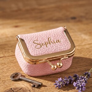 May include: A small, pink coin purse with a gold-coloured clasp and the name "Sophia" in gold script. A vintage key and sprigs of lavender are next to the purse on a wooden surface. The purse is approximately 7.6 cm wide.