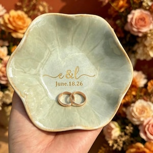 May include: A light green, petal-shaped ceramic ring dish with gold lettering that reads "e&l" and "June.18.26". Two gold wedding bands rest inside the dish. The dish is held in a hand, with a blurred floral background.