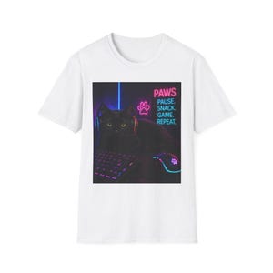 May include: White t-shirt with a graphic of a black cat wearing a headset, with a keyboard and mouse. The image includes the text "PAWS. PAUSE. SNACK. GAME. REPEAT." in pink and blue neon colours. The cat is the central focus.