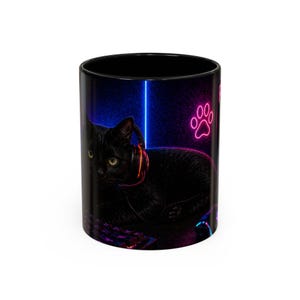 May include: Black ceramic mug with a design of a black cat wearing headphones, set against a neon blue and pink gaming backdrop. The design includes a paw print, keyboard, mouse, and a vertical blue line.
