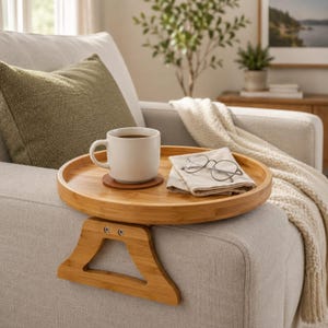 May include: A bamboo sofa arm tray with a round surface, holding a white mug of coffee on a coaster, a folded napkin, and eyeglasses. The tray has a wooden support that rests on the arm of a light-colored sofa.