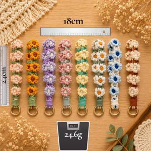 May include: A collection of handmade macrame keychains, each featuring a floral design in various colors. The keychains are approximately 2.4 cm wide and 18 cm long, with a weight of 24.6 grams. Each keychain has a gold-colored ring.