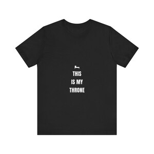 May include: A black short-sleeved t-shirt featuring a small white icon of a bed centered above the white text that reads THIS IS MY THRONE. The shirt has a crew neckline and a standard fit, with the graphic printed clearly in the center of the chest.