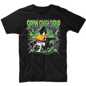 May include: Black t-shirt featuring a cartoon Daffy Duck character in a tuxedo, holding a purple drink. The shirt has the text "SIPPIN COUGH SYRUP" in green and purple, with lightning bolt accents. The design is a vibrant, cartoonish style.
