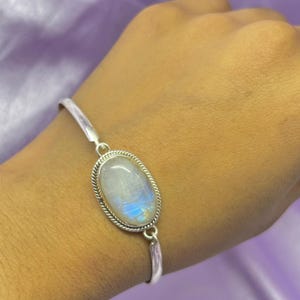 Op de afbeelding: Zilveren armband met een ovale maansteen. De maansteen heeft een parelmoerwitte basis met blauw en gele iriserende kleuren. De steen is gezet in een zilveren bezel met een gedraaid touwontwerp.