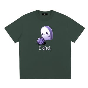 以下が含まれることがあります： 暗い緑色の半袖Tシャツ。小さな悲しそうな薄紫色の幽霊が紫色の20面ダイスを見つめているグラフィックが描かれています。グラフィックの下には、白いサンセリフ体の小文字で「I died.」というテキストがプリントされています。