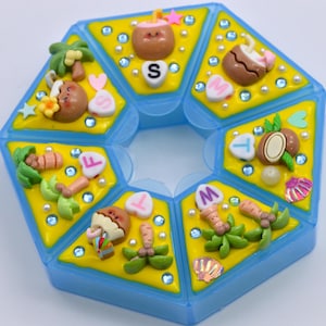 May include: A blue, multi-compartment container with yellow sections, each decorated with miniature tropical-themed embellishments. These include coconuts, palm trees, and lettered heart shapes. The container is adorned with small, clear gems.