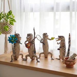 May include: Four alligator figurines in various poses. One holds blue flowers, another a guitar, a third a tennis racket and ball, and the last a small camera. A small ceramic bowl with sage sits nearby.