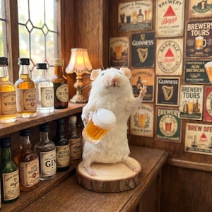 May include: A taxidermy white mouse standing on a circular wooden base, holding a miniature beer mug with foam. The mouse is positioned in front of a wall decorated with various vintage-style brewery and beer brand posters and signs.