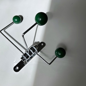 May include: A modern wall-mounted coat rack with a polished chrome finish. It features three dark green spherical knobs at the end of the arms. The rack is mounted on a rectangular chrome base with mounting holes.