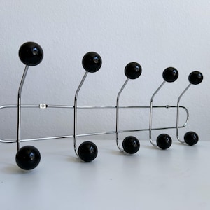 May include: A chrome metal coat rack with six black ball-shaped hooks. The hooks are evenly spaced along a horizontal bar, with a curved design at each end. The rack is designed to be mounted on a wall.