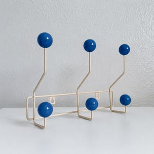 May include: A wall-mounted coat rack featuring a cream-colored metal frame with six blue spherical knobs. The rack has three higher hooks and three lower hooks, each topped with a smooth blue ball, and includes two keyhole slots for mounting.