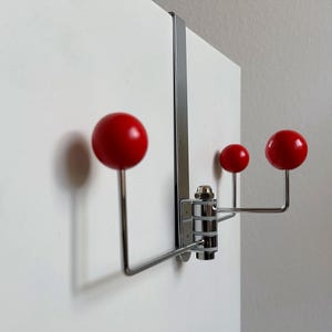 May include: A chrome-finished over-the-door hook with three red spherical knobs. The hook is mounted on a white door, with a simple, modern design. The knobs are evenly spaced and add a pop of color.