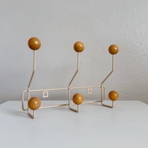 Puede incluir: Un perchero de pared con estructura de metal beige claro y seis ganchos redondeados. Cada gancho está rematado con una esfera de madera lisa de color amarillo mostaza. El marco incluye dos ranuras integradas para el montaje en la pared.