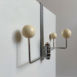 May include: A chrome-finished over-the-door hook with three cream-colored ball-shaped knobs. The hook is designed to hang over a door and has a modern, minimalist aesthetic. The knobs are evenly spaced.