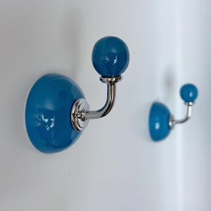 Könnte beinhalten: Zwei blaue Keramik-Wandhaken mit glänzendem Finish. Jeder Haken hat eine abgerundete Basis und einen kugelförmigen Knauf, verbunden durch einen gebogenen, silberfarbenen Metallarm. Die Haken sind an einer weißen Wand befestigt.