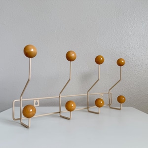 May include: A mid-century modern coat rack with a cream-colored metal frame and six wooden ball-shaped knobs. The knobs are a warm, amber color. The rack is designed with a horizontal bar and angled hooks.