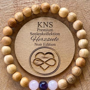 Op de afbeelding: Een ronde houten plaat gegraveerd met KNS Premium Seelenkollektion Herzseele Noir Edition en een oneindigheidshart-symbool, omringd door een armband van lichtbruine houten kralen, twee lichtroze kralen en één donkerpaarse kraal.