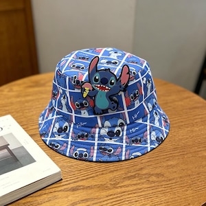 Puede incluir: Sombrero de pescador azul con un patrón repetido del personaje Stitch de Lilo & Stitch, con una aplicación 3D de Stitch en la parte delantera. El sombrero tiene un diseño de cuadrícula y la palabra "Hi".