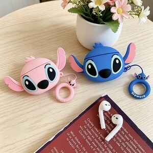 May include: Pink and blue cartoon character-shaped AirPod cases with matching keychains. The cases have large eyes and ears. A pair of white AirPods and a book are also visible.