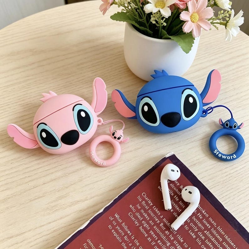 May include: Pink and blue cartoon character-shaped AirPod cases with matching keychains. The cases have large eyes and ears. A pair of white AirPods and a book are also visible.