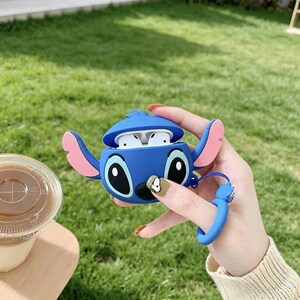 May include: A blue silicone case shaped like a cartoon character, with large black eyes and pink ears, holding white earbuds. The case has a blue wrist strap. The background is green grass.