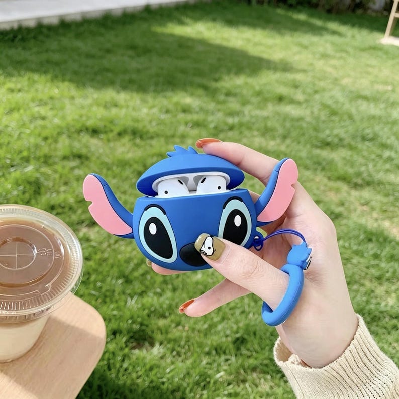 May include: A blue silicone case shaped like a cartoon character, with large black eyes and pink ears, holding white earbuds. The case has a blue wrist strap. The background is green grass.