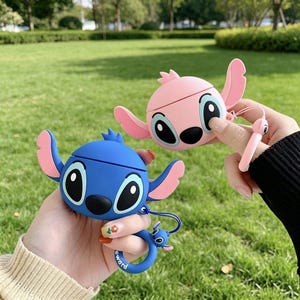 May include: Two cartoon character-themed AirPod cases. One is blue with pink ears, the other is pink. Both have large eyes and are held up against a green background. The blue case has a blue ring with a small character attached.