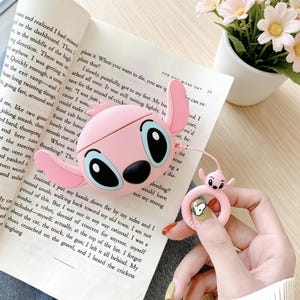 May include: Pink silicone AirPod case and ring holder featuring a cartoon character design. The case is shaped like the character's head with large eyes and ears. The ring holder has a small figure of the character.