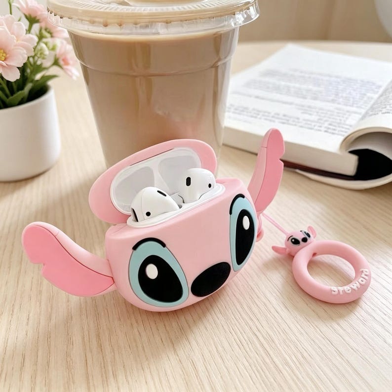 May include: Pink silicone AirPod case shaped like a cartoon character with large black eyes and blue accents. The case is open, revealing white earbuds. A matching pink ring with a small character charm is also present.