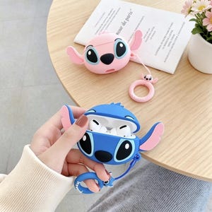 May include: Two Stitch-themed AirPod cases in pink and blue. The blue case is open, revealing white earbuds. Both cases have large black eyes, pink ears, and a matching keychain. A pink ring-shaped keychain is also visible.