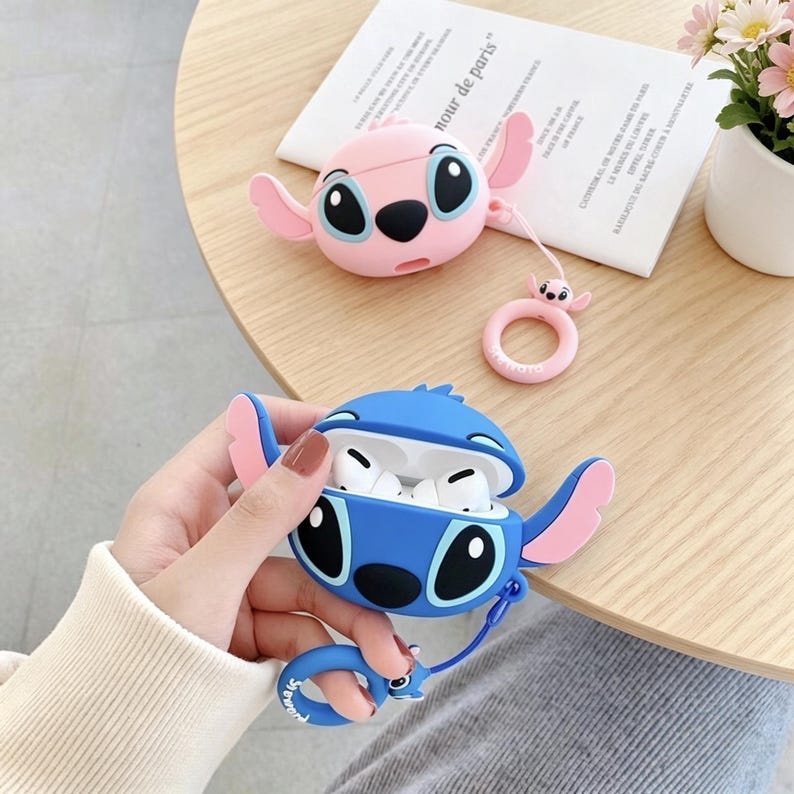 May include: Two Stitch-themed AirPod cases in pink and blue. The blue case is open, revealing white earbuds. Both cases have large black eyes, pink ears, and a matching keychain. A pink ring-shaped keychain is also visible.