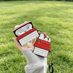 Puede incluir: Una funda para AirPods roja, blanca y negra con el tema de Snoopy, abierta para revelar auriculares blancos. La funda tiene la forma de la caseta de Snoopy, con una figura de Snoopy a juego en el llavero adjunto. El fondo es hierba verde.
