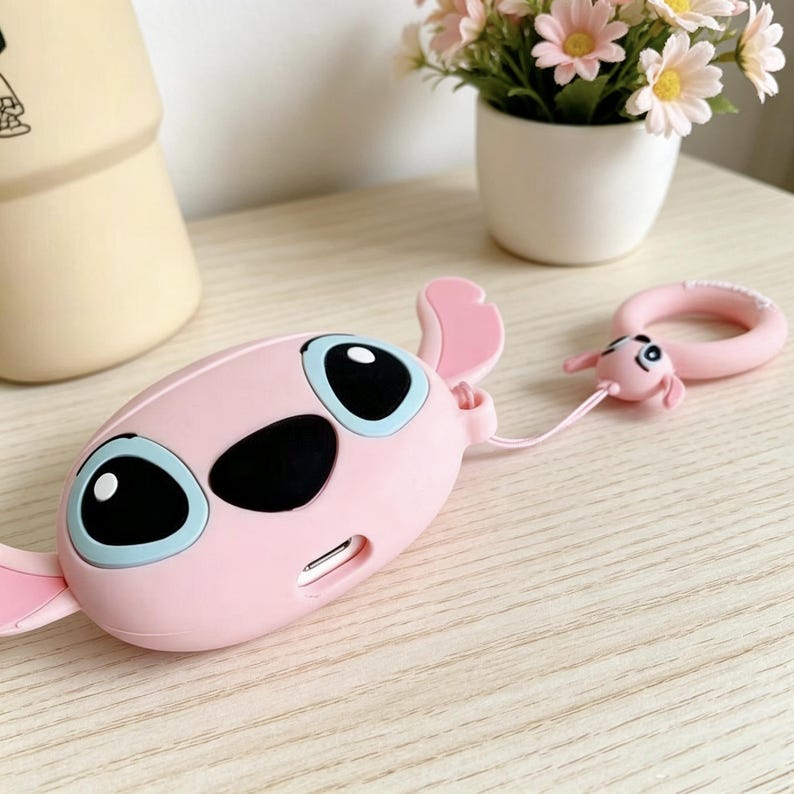 May include: A pink, cartoon-style case with large black eyes and blue accents. The case has a matching pink keychain with a small character figure. The design is reminiscent of a popular animated character.