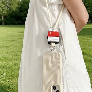 Puede incluir: Una funda de teléfono color crema con un cordón temático de Snoopy. El cordón presenta un dije de casa de Snoopy con detalles rojos y blancos. La funda tiene una superficie texturizada y un anillo plateado.