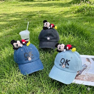 May include: Three denim baseball caps with Mickey Mouse plush toys. The caps are in shades of blue and grey, with the text "OK Be Luck" and a Mickey Mouse graphic. The caps are on green grass.