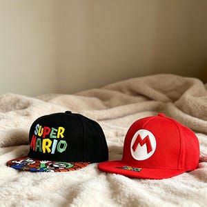 Op de afbeelding: Twee baseballpetten met platte klep. Eén is zwart met "SUPER MARIO" geborduurd in kleurrijke letters en een patroon op de klep. De andere is rood met een wit "M"-logo en een bijpassende klep.