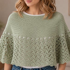 May include: A sage green crochet poncho featuring a solid, tightly stitched yoke and a wide, flared lower section with an intricate, openwork fan pattern. The garment has a round neckline and a scalloped hemline, worn over a plain white shirt.