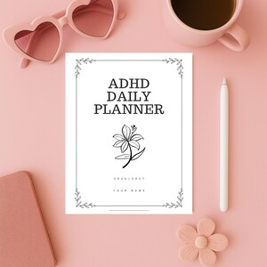 Op de afbeelding: Een witte ADHD Daily Planner met een bloemenillustratie en de tekst "ADHD DAILY PLANNER" in het zwart. De planner bevat de jaren 2026/2027 en een ruimte voor een naam. Roze hartvormige zonnebrillen, een roze mok en een witte pen zijn ook zichtbaar.