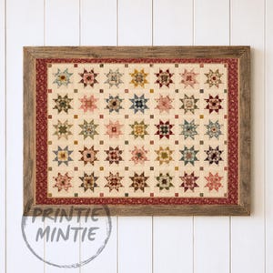 Op de afbeelding: Een ingelijste quilt met een raster van veelkleurige sterpatronen op een crèmekleurige achtergrond, omrand door een donkerrode bloemenstof. De lijst is gemaakt van verweerd bruin hout. De tekst PRINTIE MINTIE is linksonder zichtbaar.