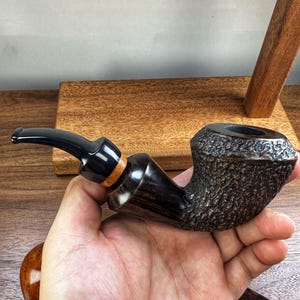Op de afbeelding: Een tabakspijp met een donkerbruine, getextureerde, rustieke kop en een gladde, gepolijste zwarte steel. Het mondstuk is zwart met een dunne, lichtgekleurde houten accentring bij de verbinding, wat zorgt voor een contrast in textuur.