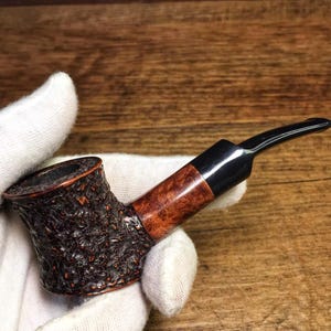 Pode incluir: Um cachimbo de tabaco com um fornilho rústico de cor castanho-escuro e textura profunda, que transita para um canudo liso de cor castanho-avermelhado polido e uma piteira preta de acrílico. O cachimbo tem uma forma curva e uma extremidade larga.