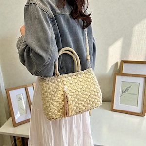 May include: A woven straw handbag with a rectangular shape, featuring a braided handle and a long, detachable shoulder strap. The bag has a decorative tassel and is light beige in color.