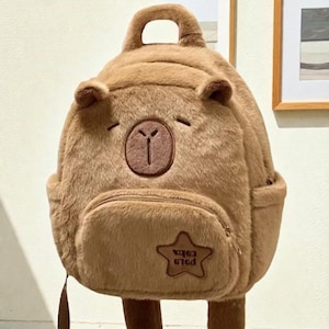 May include: Brown plush backpack shaped like a bear with a small top handle and adjustable straps. The backpack features a front zippered pocket with a star-shaped patch that has the text "x463 616d". The bear's face is embroidered with closed eyes and a circular emblem.