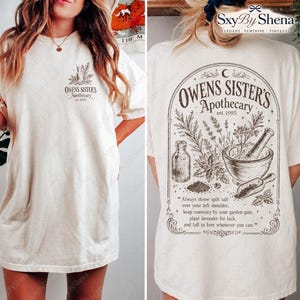 May include: An off-white t-shirt featuring a small apothecary-themed graphic on the front left chest and a large, detailed back print. The back design displays a mortar and pestle, herbs, and the text: Owens Sister's Apothecary est. 1995.