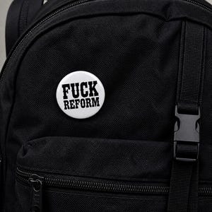 May include: A black rucksack with a white button pinned to it. The button displays the words "FUCK REFORM" in bold, black capital letters. The rucksack has multiple compartments and a black buckle.